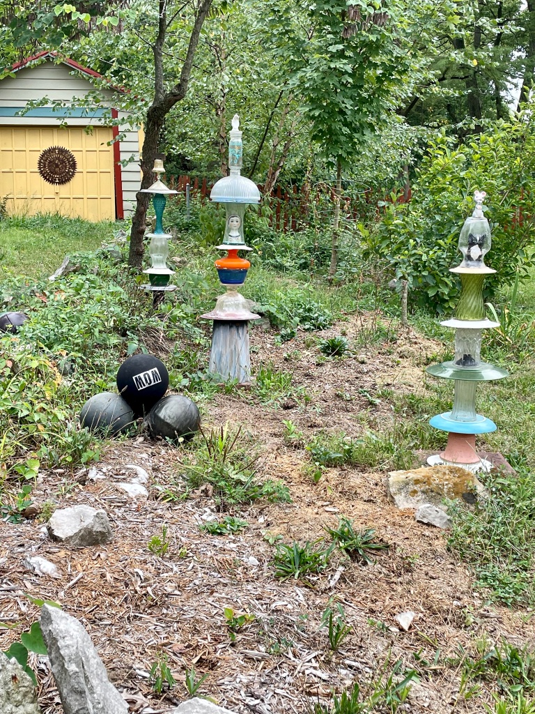 Yard with three small tower-like objects constructed from a variety of found objects, many of them glass vases or lamp shades. Some of the vases contain objects - doll head, ceramic cat. In background is a detached garage colorfully painted with a sunburst design on the door. Three large black balls are stacked pyramid style in the foreground.
